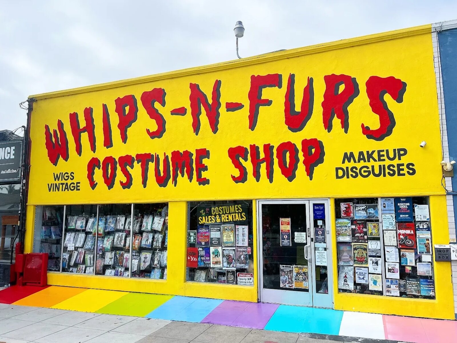 The bright yellow Whips 'n Furs Costume Shop storefront on 30th Street, North Park, with its hand-painted dripping red sign reading Costume Shop, Wigs, Vintage, Makeup, Disguises, above a rainbow-painted sidewalk