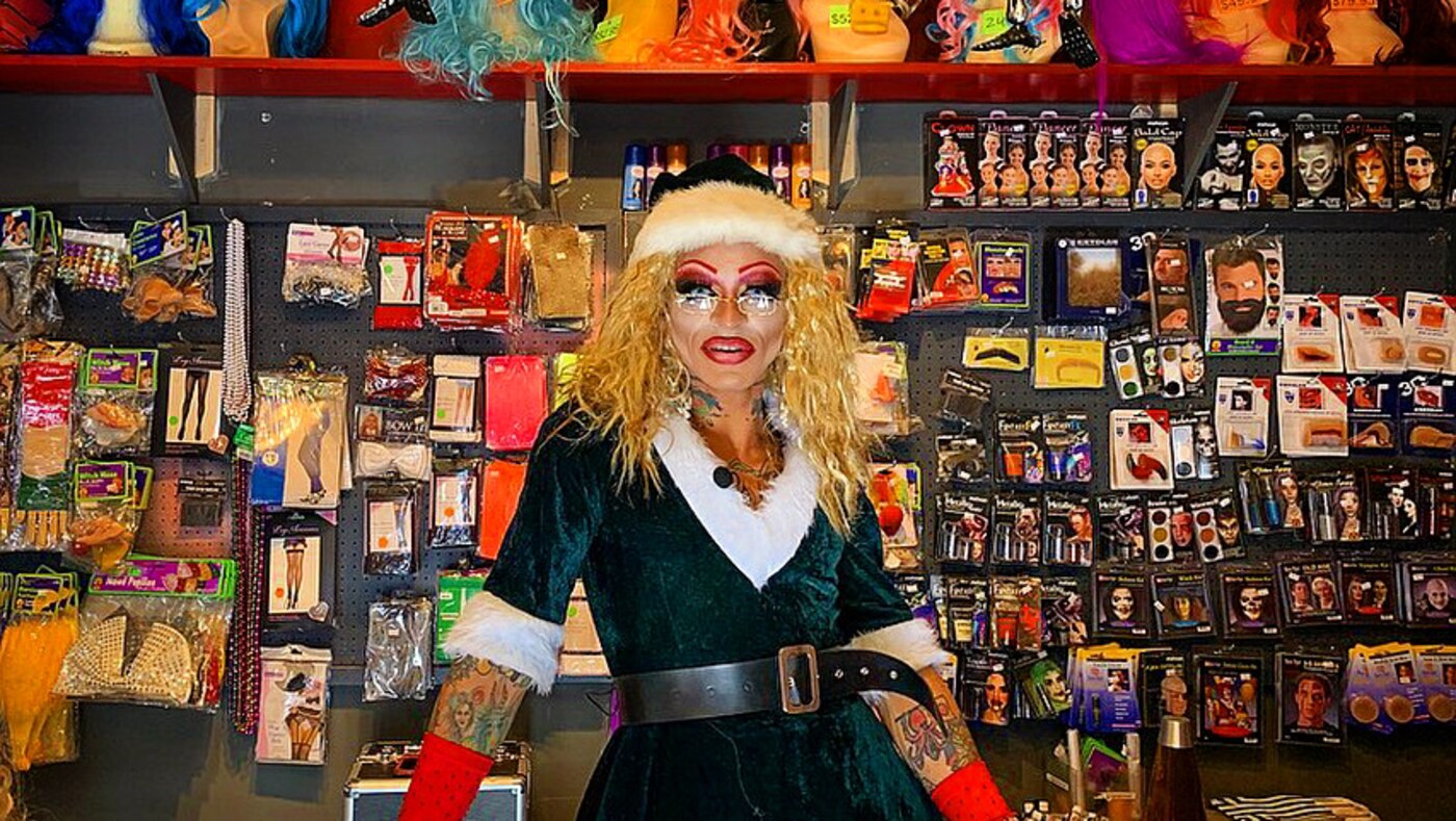 A person in full glam and SFX makeup and a blonde wig standing in front of the theatrical makeup wall at Whips 'n Furs, lined with Ben Nye, Kryolan and Mehron kits